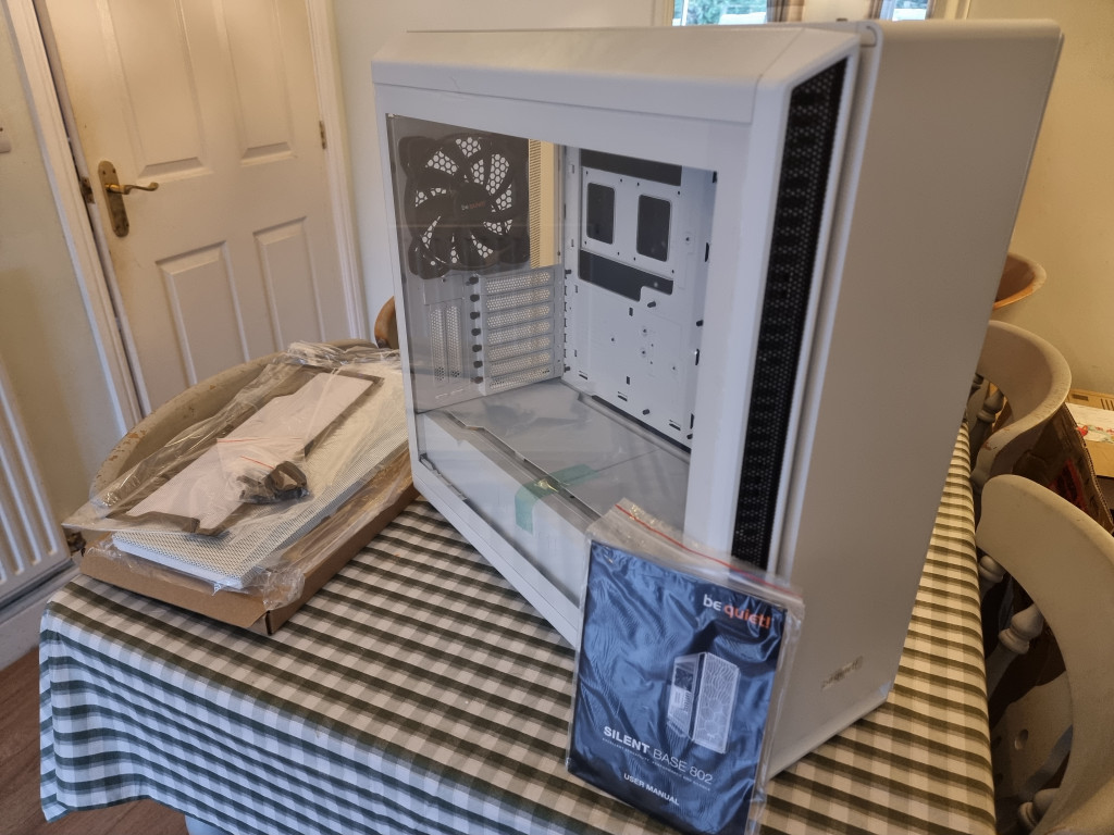 A large be quiet! Silent Base 802 PC case stands on a dining room table. It has a glass window side that looks into the vast space within. An alternative mesh front cover lies inside a carboard box to the left while a manual is propped up infront of the PC case.