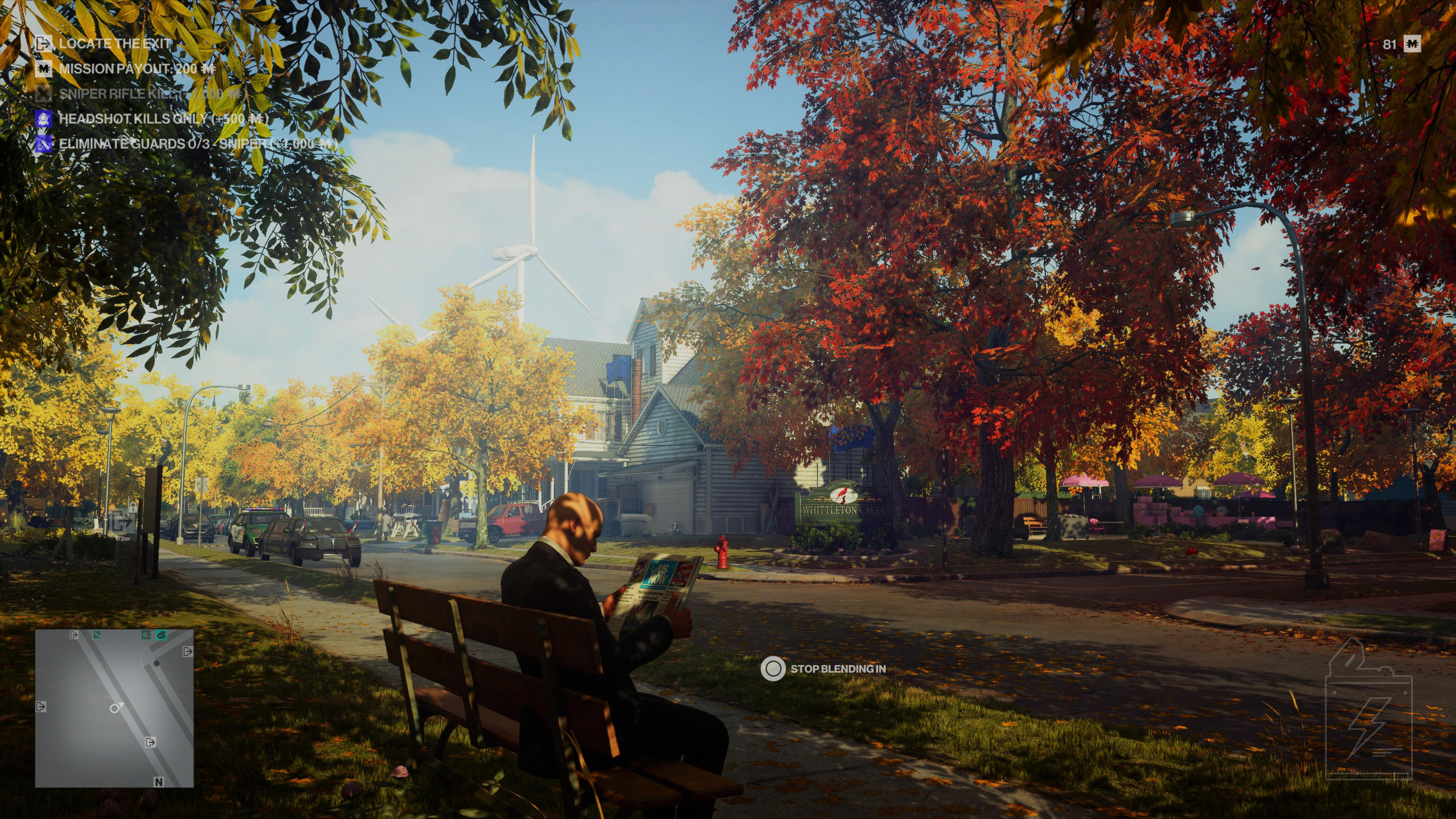 Agent 47 sits on a park bench in a on a suburban street in a residential area reading a newspaper in order to blend in during a Hitman Freelancer mission. It is autumn time as the trees around have various colours of red yellow and orange foliage.
