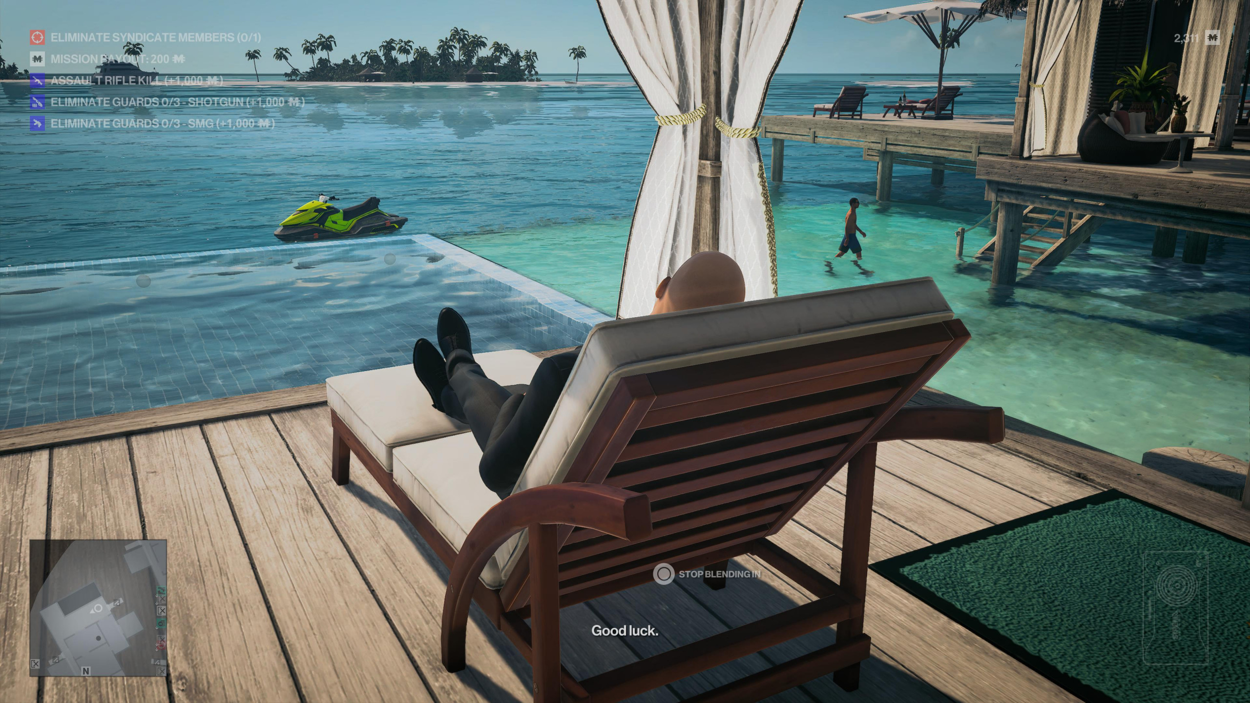 Agent 47 relaxes on a sun lounger in a tropical paradise with turquoise waters in front of him. There are palm trees and sand in the distance