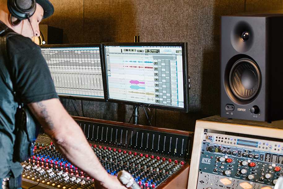 image showing a music producer infront of a mixing desk with 2 monitor screens above. To the right is a black Edifier MR4 studio monitor speaker