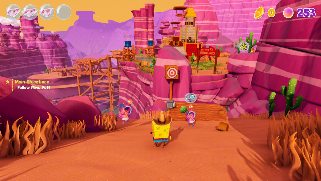 Screenshot showing spongebob in wild west costume following mrs puff along a dirt track adourned with brown sea grass and green cacti. The scenery is a pink and purple canyon featuring numerous wooden platforms and bridges. the sky is yellow.