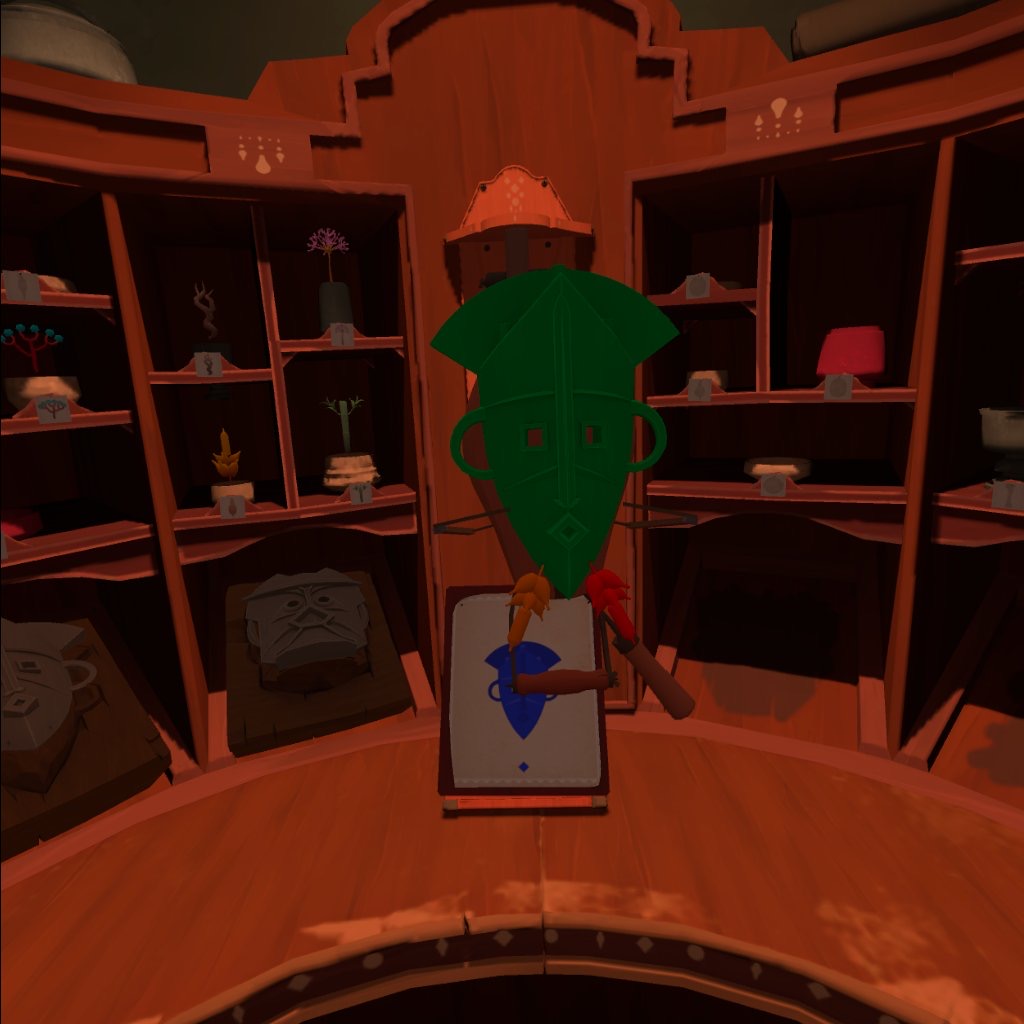 A green wooden mask rests on a wooden stand. Unusual objects stick out of it's chin. Behind are boxes full of mask components. Below the mask is a template with a plain blue mask on it. 