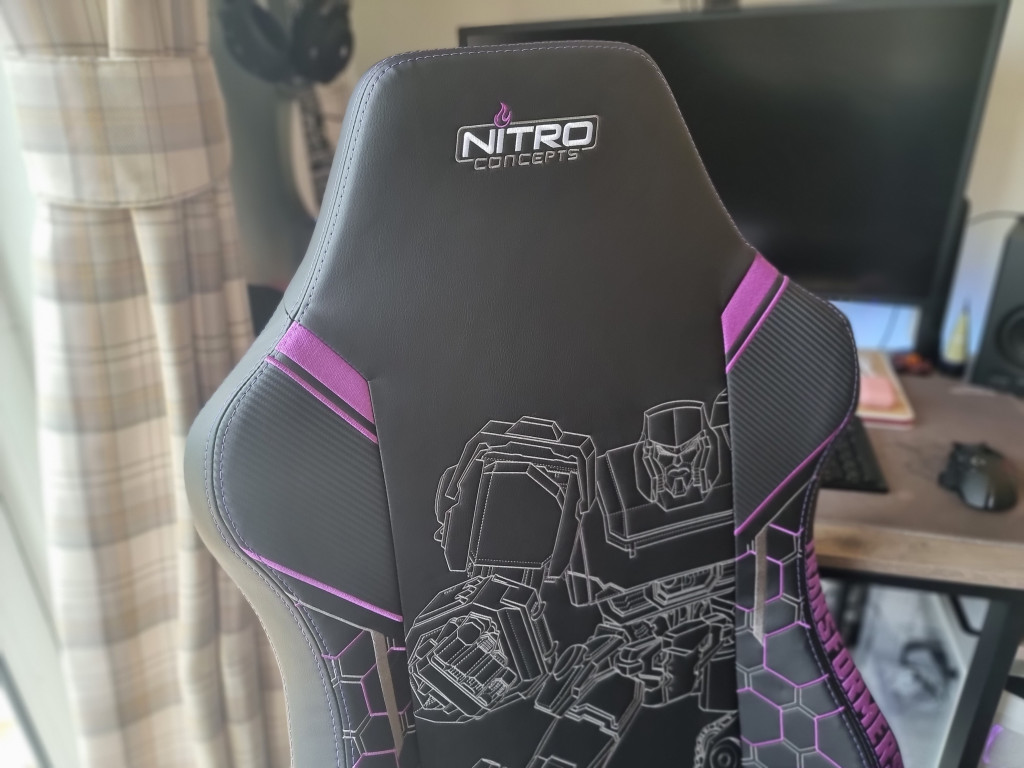 picture showing the top section of the seat back with the logo "Nitro Concepts" on the headrest in white, silver and purple. Further down the chair you can see the silver stitched outline decal of Megatron as well as more purple accents.