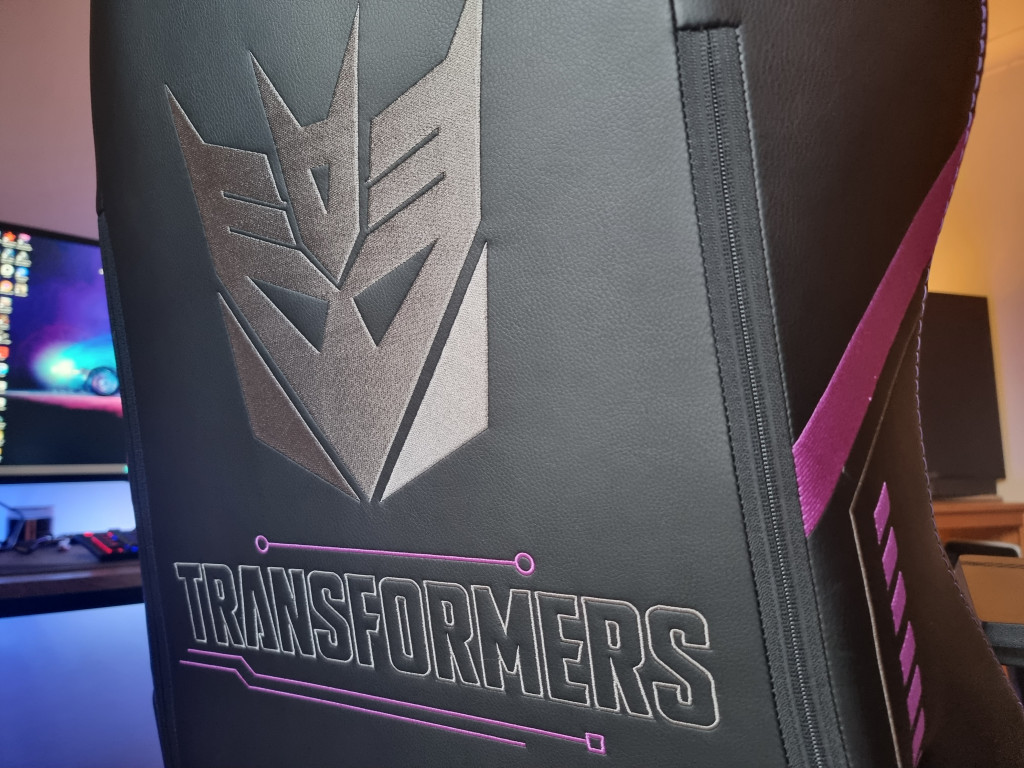 picture showing the rear of the black gaming chair with a large stitched silver Decepticon logo in the middle. Below it is the outline of the word "Transformers" along with purple lines above and below.