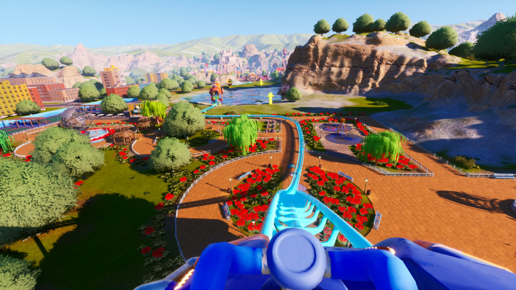 screenshot showing a first person view at the front of my blue roller coaster train. The light blue track is sweeping downwards towards the ground through a orange paved area surrounded by red flowers and green trees. In the background is a blue lake and a cliff line along the right hand side.