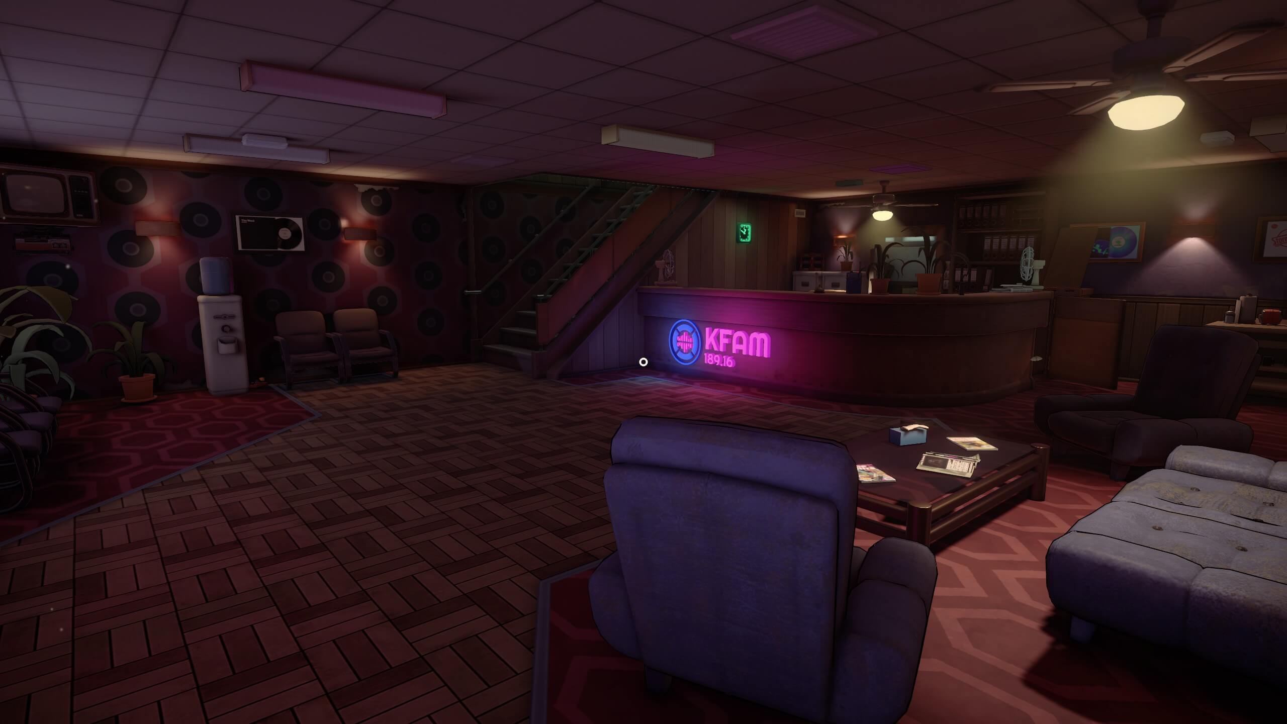 the downstairs area reception of KFAM, showing a set of chairs to the right and the reception desk near the glowing neon. The stairs leading up to the main recording area to the top of the screen. 