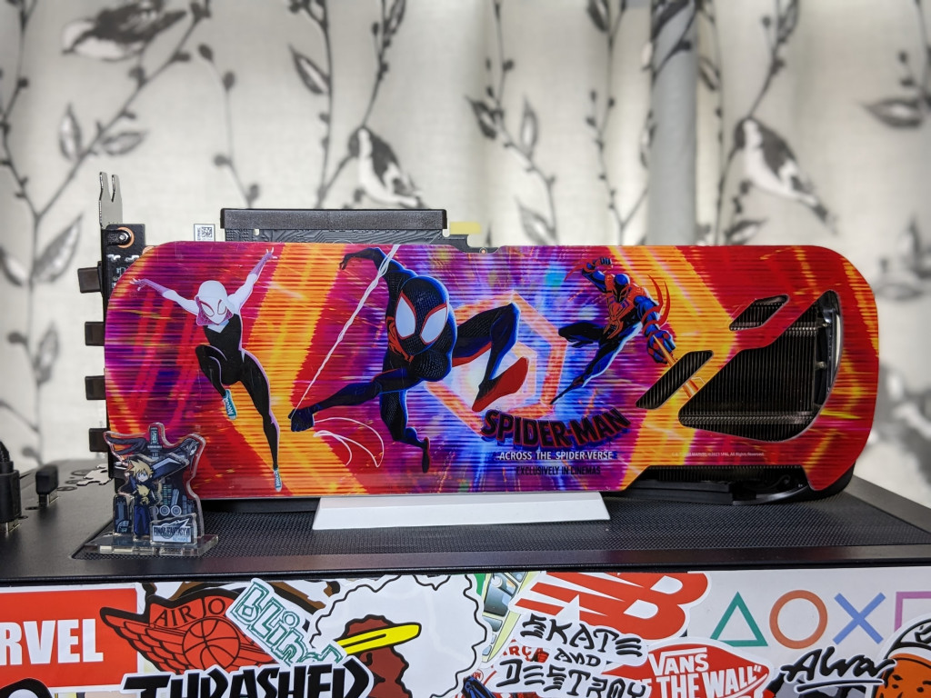 Shot of the backplate of the RTX 4070 Ti showing, a scene from the movie Spider-Man, across the Spider-Verse, with Spider-Gwen, Miles Spider-Man and Spider-Man 2099 from left to right. THe GPU is mounted on top of a PC that has a sticker bombed back panel. In front of the GPU is a little plastic stand showing Shinra Tower, Sephiroth and Cloud from Final Fantasy 7 Remake. In the background are grey curtains with birds on.
