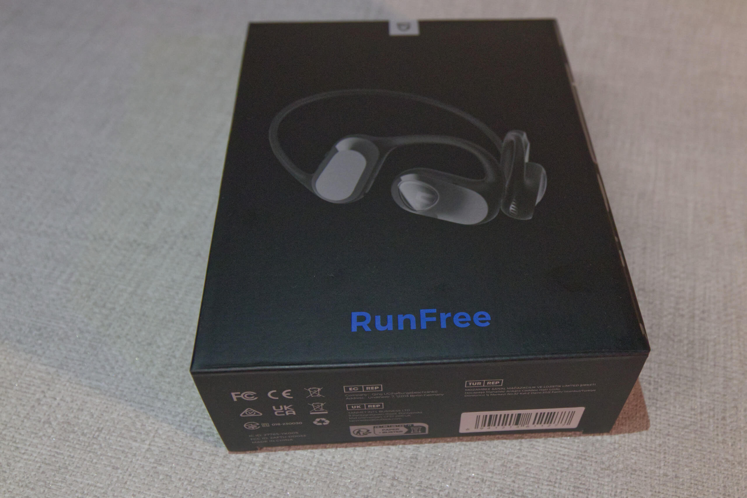 Photo shows the SoundPeats RunFree headphones in their original packaging/box. The box is a dark grey with blue logo, and the earphones are pictured in the centre