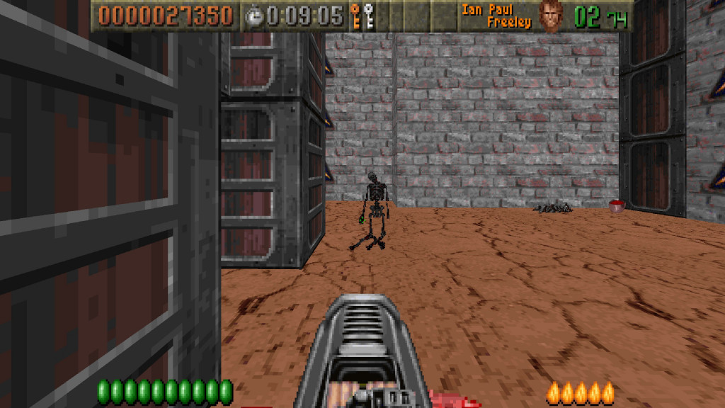 screenshot showing a flamewall gun having been recently used. Infront is the charred remains of a black skeleton that is still standing. The floor is light brown and cracked and the walls are a mixture of stone and metal blocks.