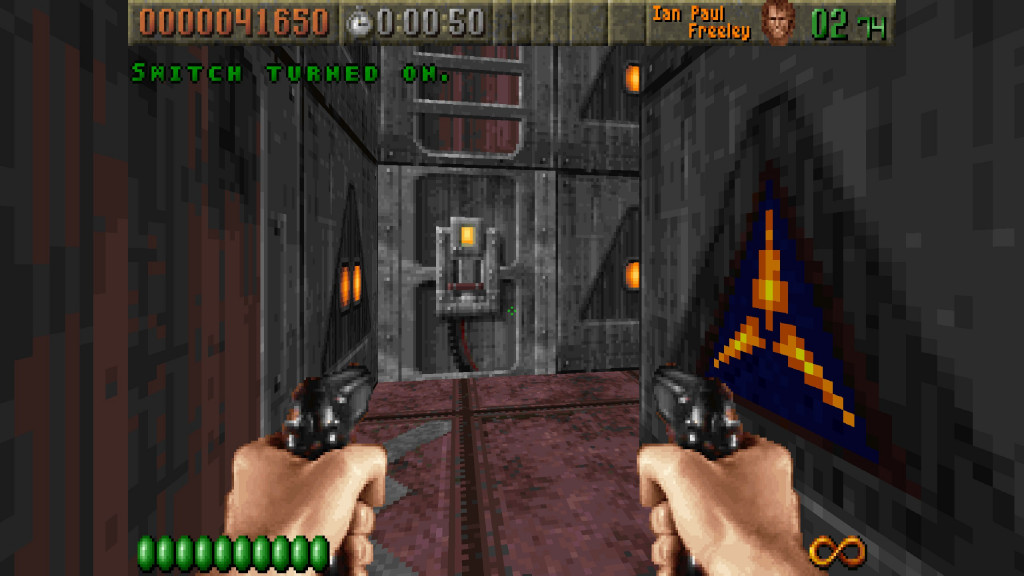 screenshot showing an fps view with dual pistols as the weapon in use. Infront is a switch to trigger a door. The floor is pink slabs and the high walls behind are grey metal looking blocks with a yellow triad logo.