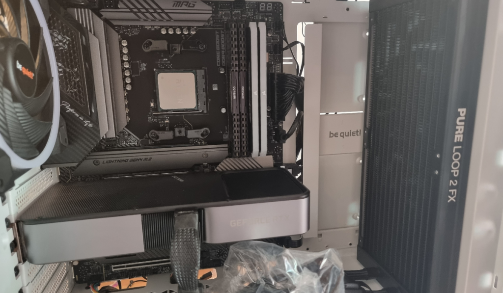 photo showing the inside of the my white PC case. The dark grey MSI motherboard can be seen in the background with the shiny silver Ryzen 5 3600 CPU awaiting the installation of the cooler block. Visible is the RTX 3070 GPU and 4x8GB DDR4 Ram. The black Radiator is propped up vertically towards the front of the case and a bag of screws lies in the bottom.