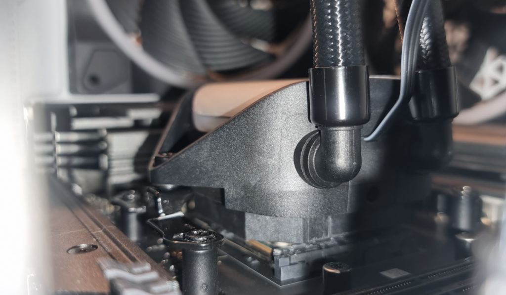 photo showing a super close up of the be quiet cpu cooler block installed over the CPU socket. The camera is low against the motherboard, staring at the side of the black cpu cooler block. A black radiator hose can be seen going away from it vertically. In the background is a 140mm light wing exhaust fan.