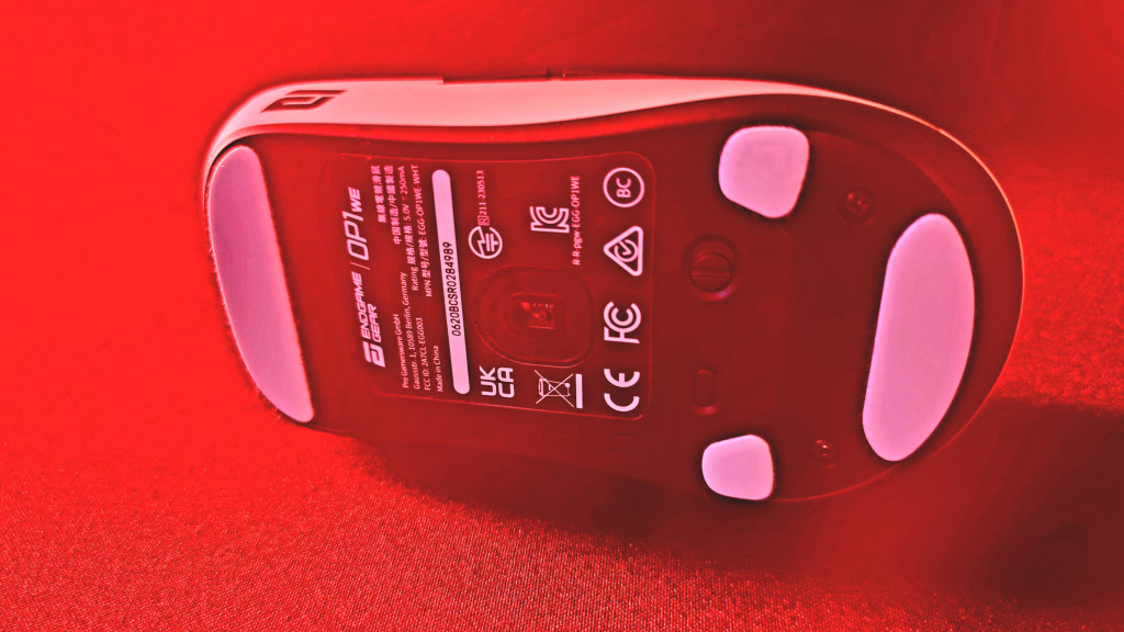 photo of the white endgame gear gaming mouse on its side with the camera looking at the black underside of it. The white skates that the mouse glides on can be seen. 1 at the front and 3 at the back. The mouse is bathed in red light.