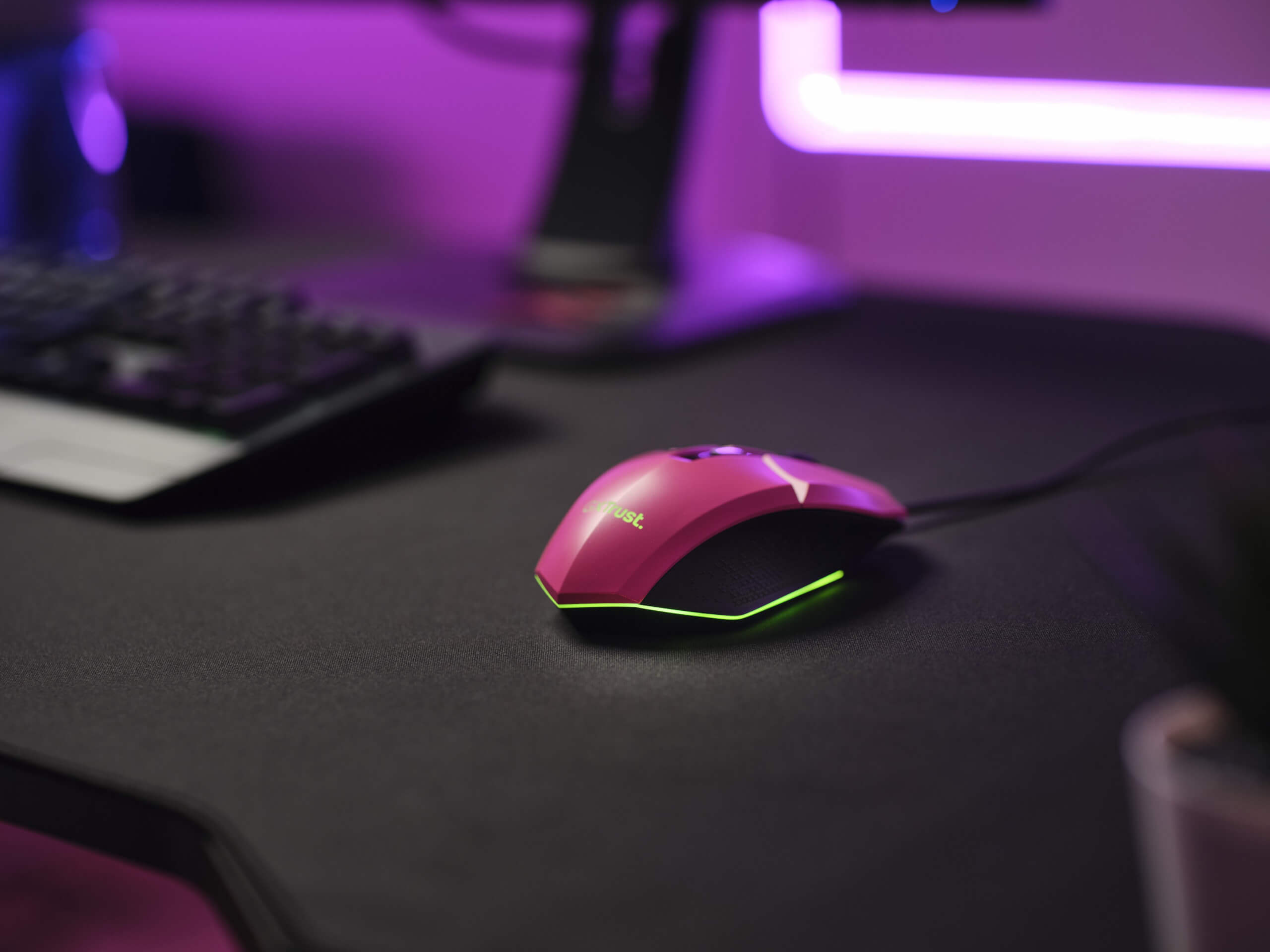 image showing the trust gaming mouse on a desktop. With pink colouring and a green rgb strip along the bottom of the mouse. in the background is a keyboard and other neon lights.