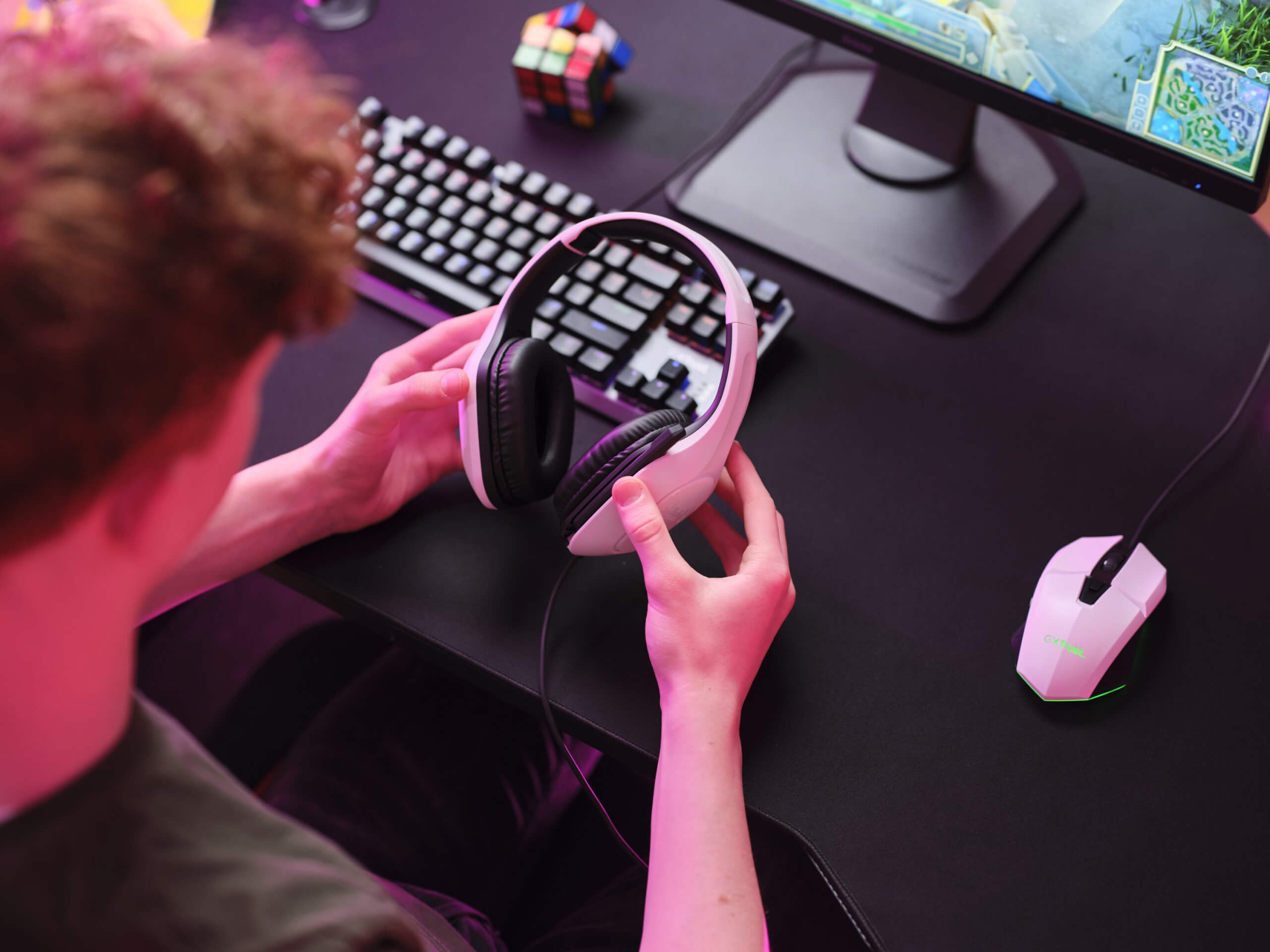 a headset being held in the hands of a gamer at a desk. With rubiks cube, keyboard and mouse on screen