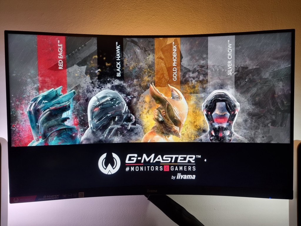 photo showing the monitor mounted to my wall with it's trademark iiyama gaming wallpaper. The image features 4 characters, representing the 4 different series of hardware that they manufacture. Red eagle, black hawk, gold phoenix and silver crow.