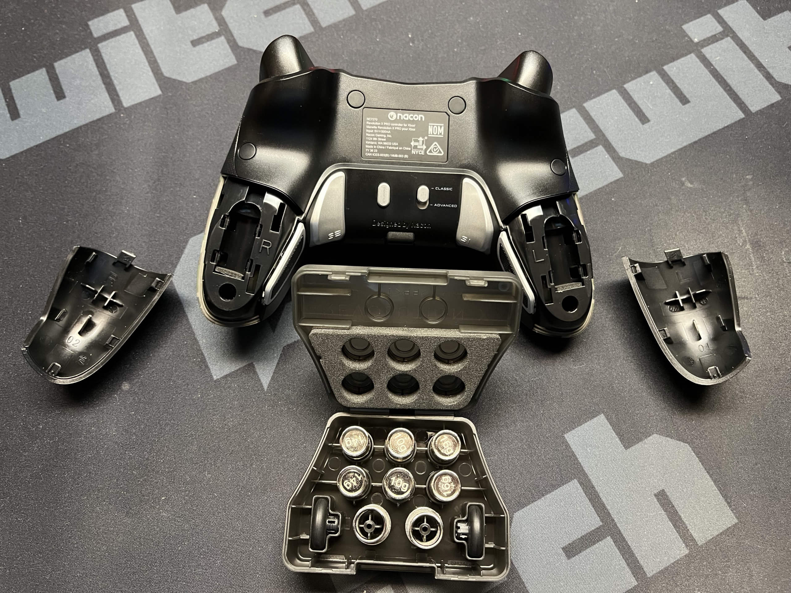 Weights are exchanged on the back side of the Revolution X Pro. The back side of the controller with the two handhold parts open. A small box with the exchangeable weights, thumbsticks, and posts is shown as well.