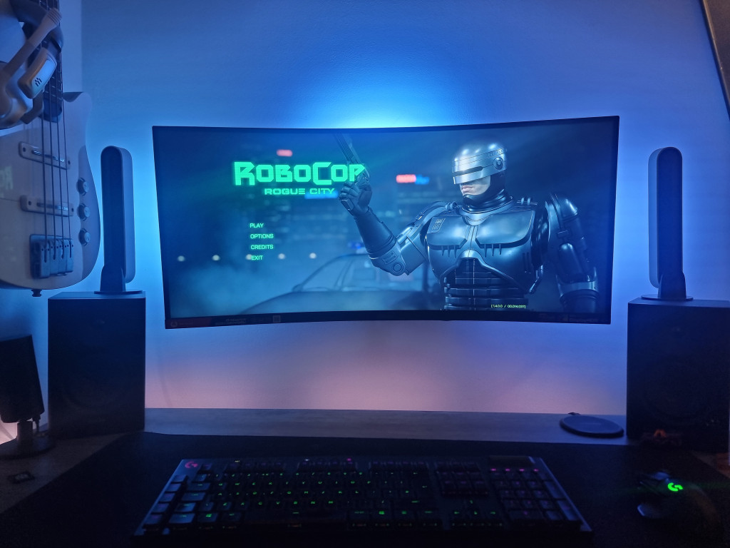 photo showing the monitor on the wall with no wires shown. Robocop is displayed and the wall is lit in blue light behind the screen. The hue play bars are visible on the black speakers either side while a white bass guitar hangs from the left wall.