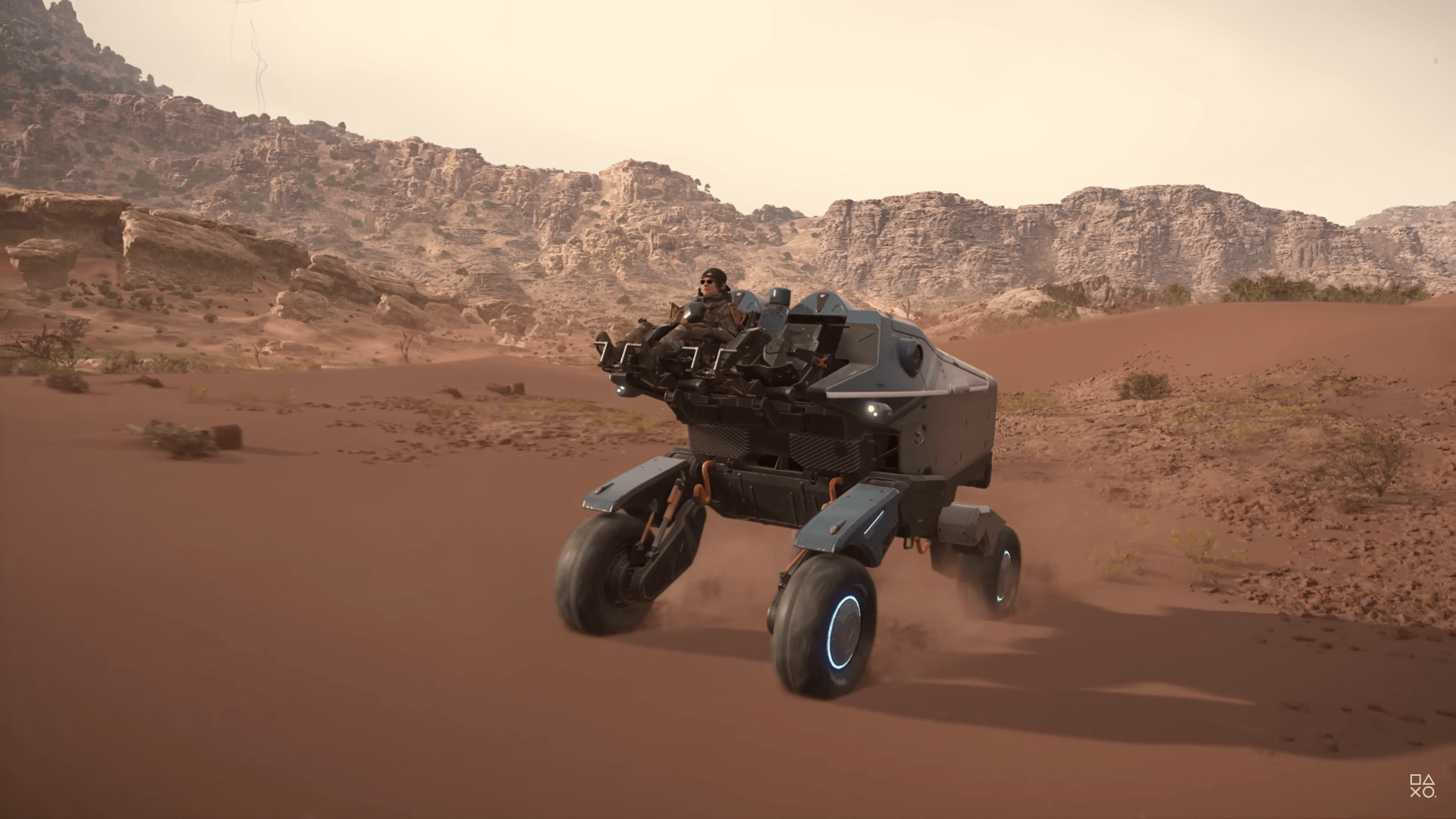 a guy wearing sunglasses rides an open fronted 2 seater rover vehicle across desert terrain with rocky areas in the background