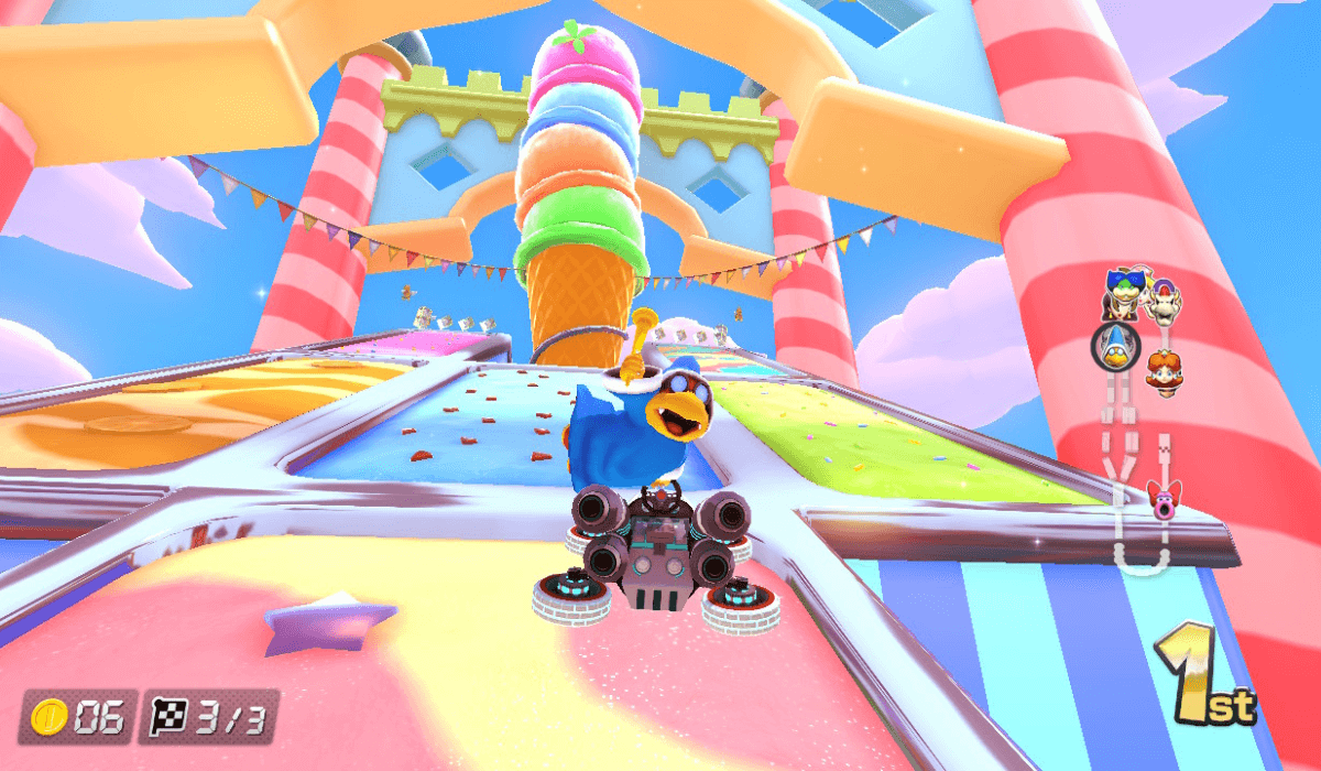Kamek, a new Mario Kart character, floating above his Go Kart mid-race and posing for the camera with a flourishing wand. The track behind him is made of candy and sweets, and he is in first place.