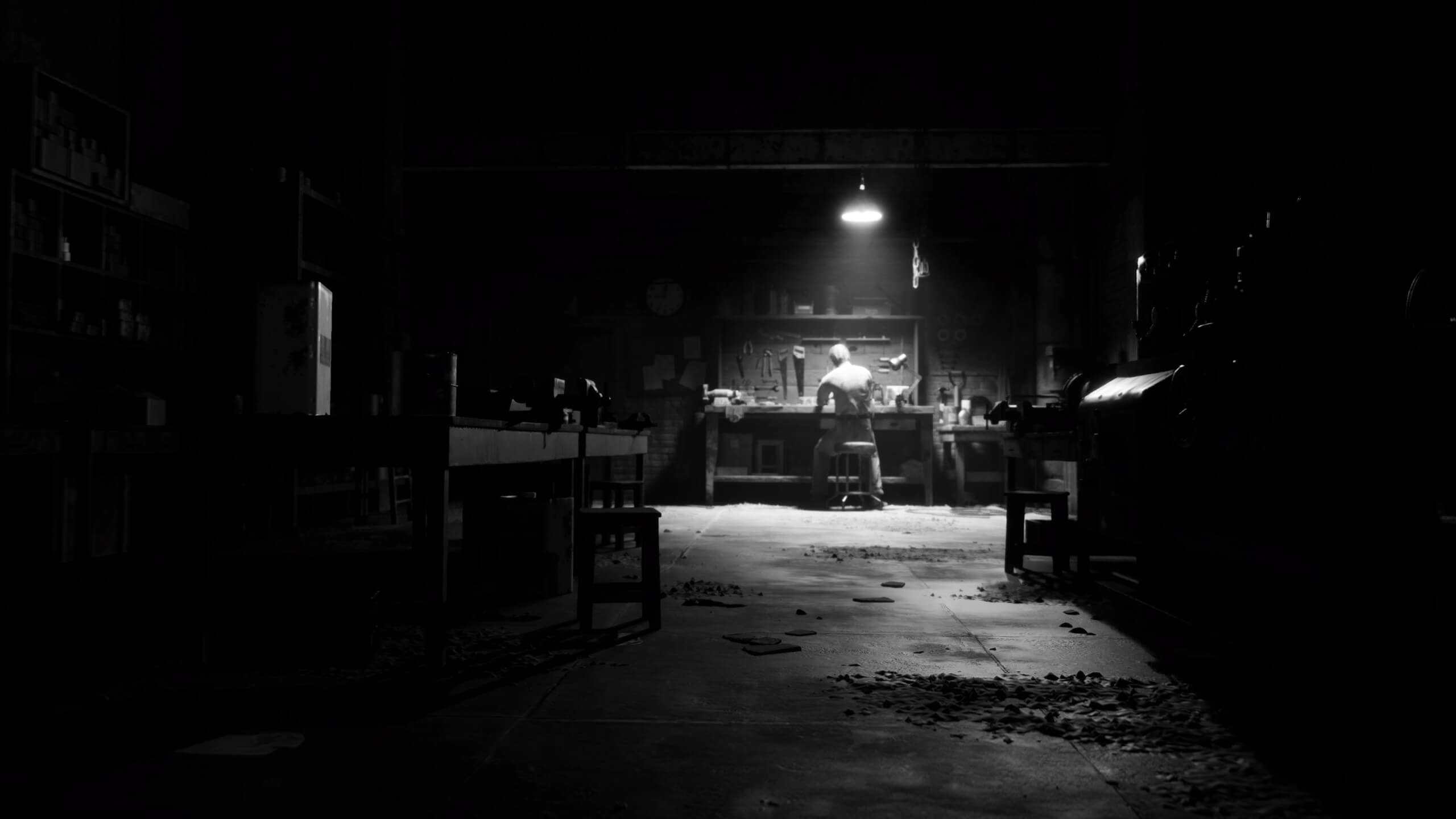 A lone man is sitting in his work shop. A light beams down on him to add more of a menacing visual. The whole shot is covered with a black and white filter.