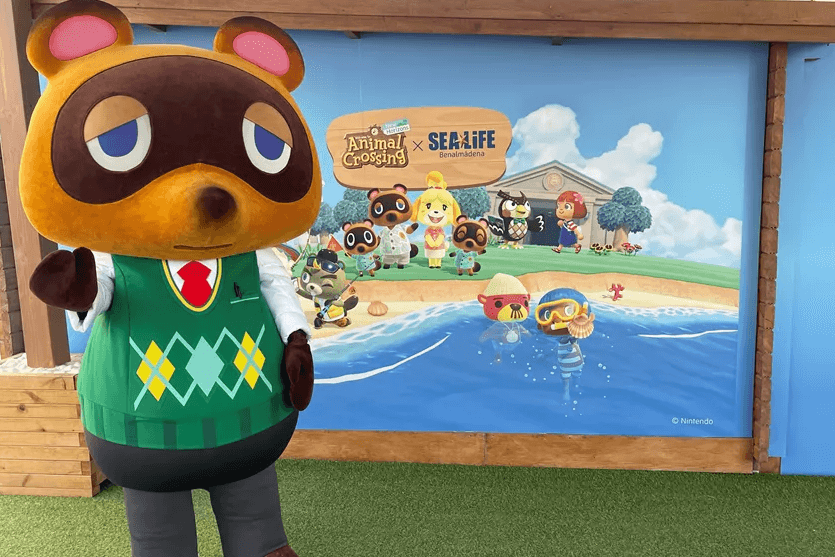 A character suit Tom Nook waves whilst standing in front of a Sea Life Animal Crossing: New Horizons event sign