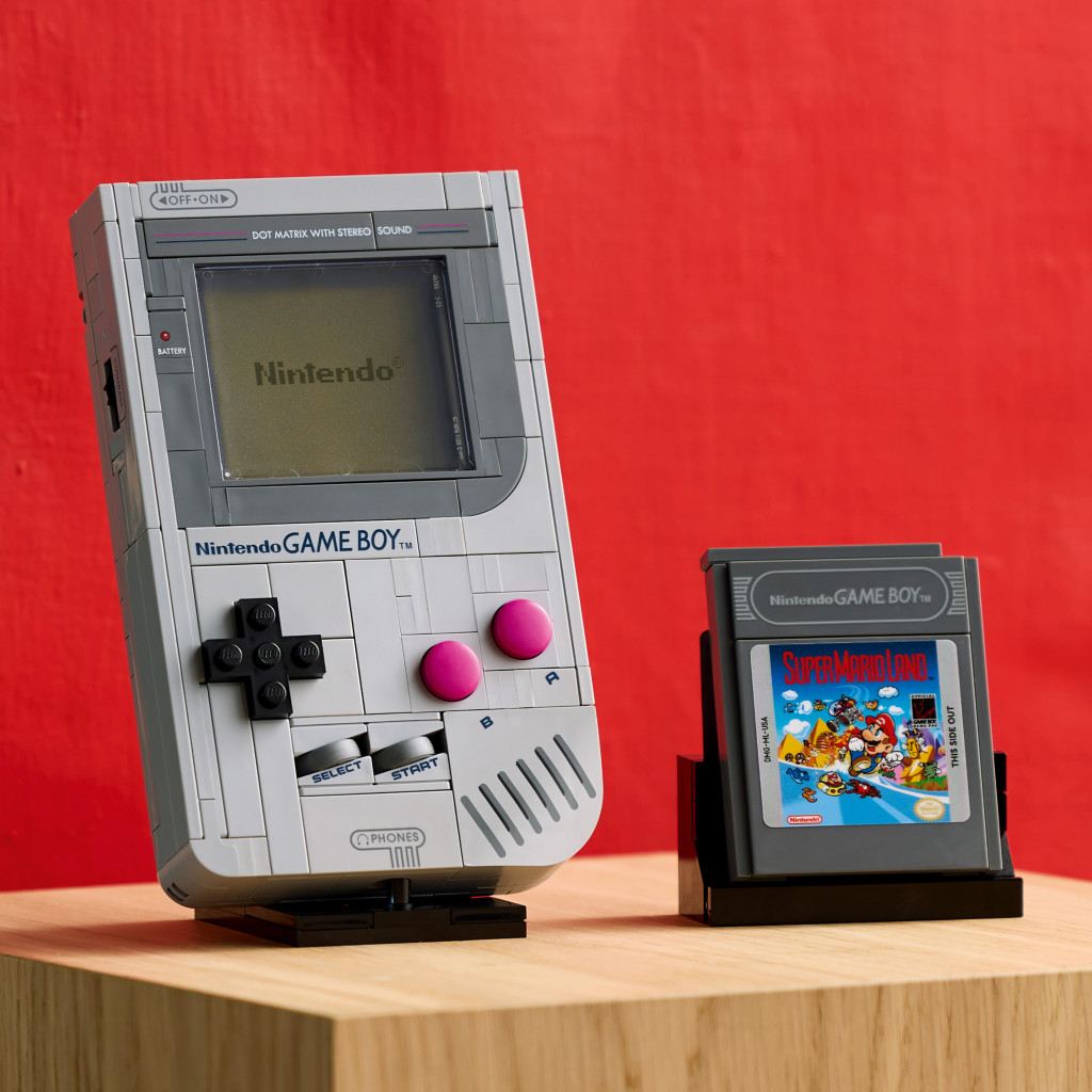 A Nintendo Game Boy console built from LEGO sits on a wooden stand, next to the game cartridge for Super Mario Land, against a red background.