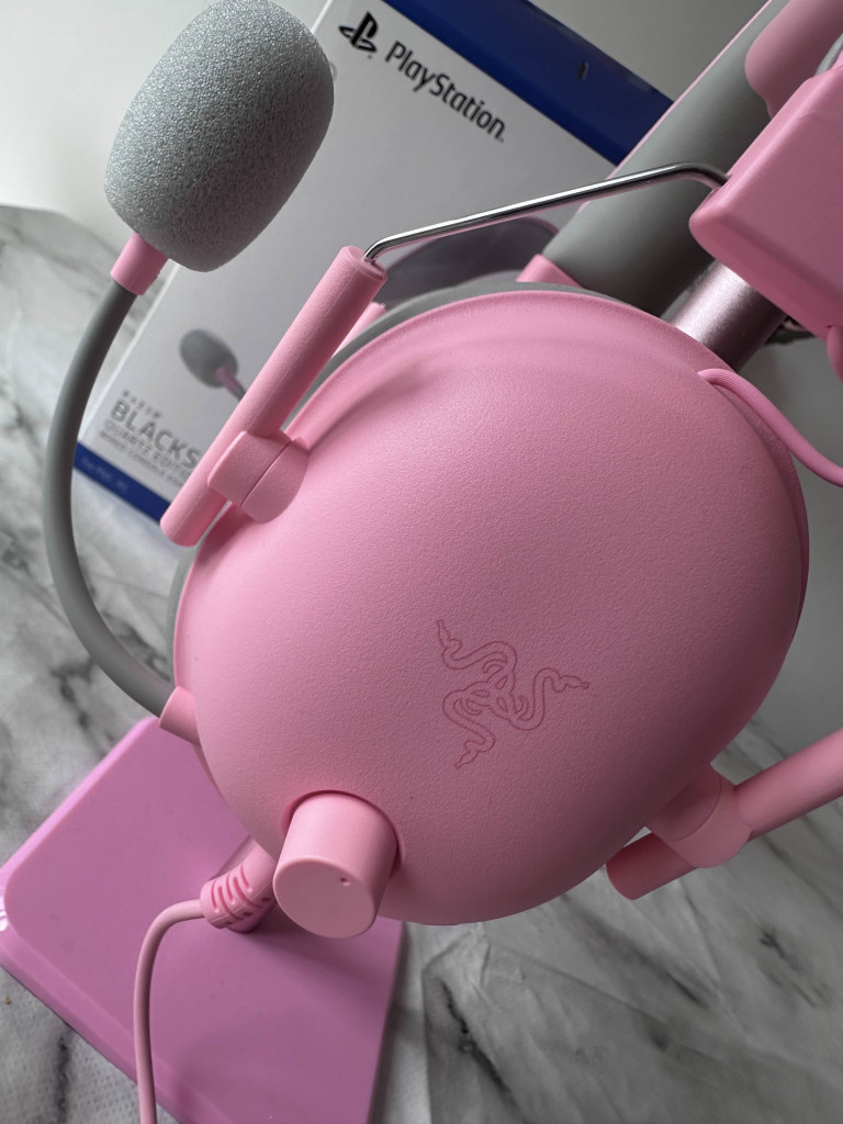 Close-up of a Pink Razer BlackShark V2 X headset with a microphone, resting on a matching base, against a PlayStation background.