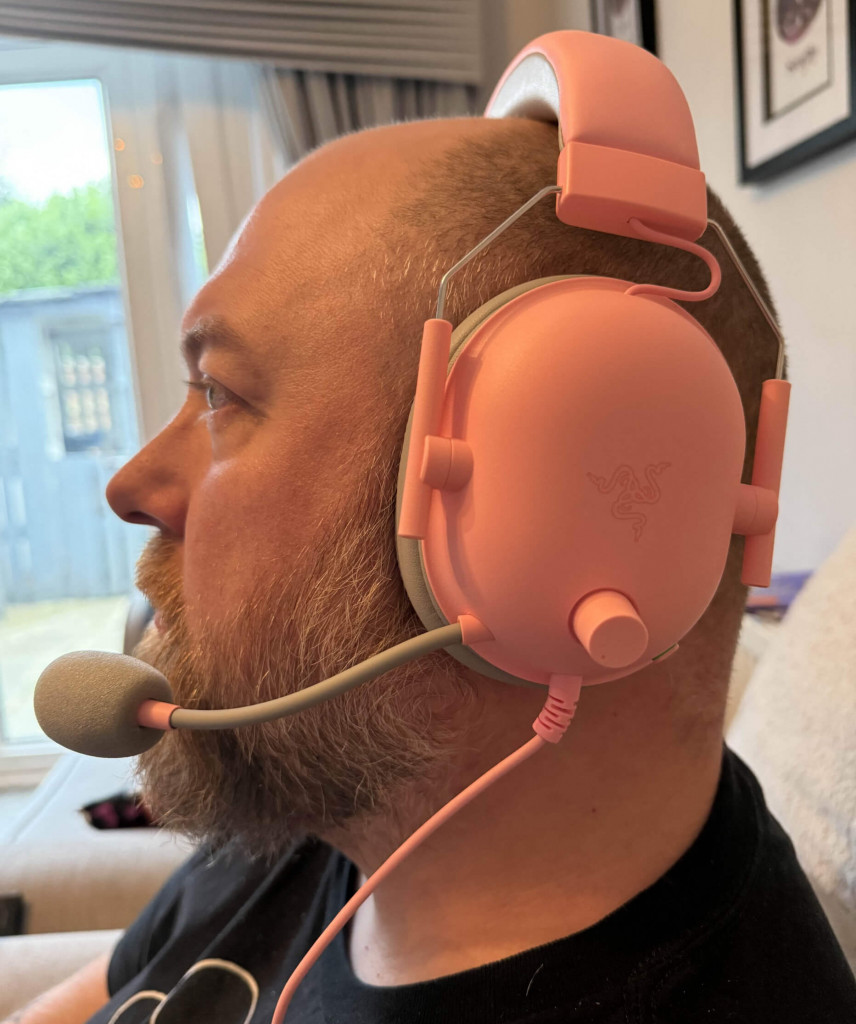 A person is wearing pink gaming headphones with a microphone, sitting comfortably on a couch in a cosy, well-lit room.