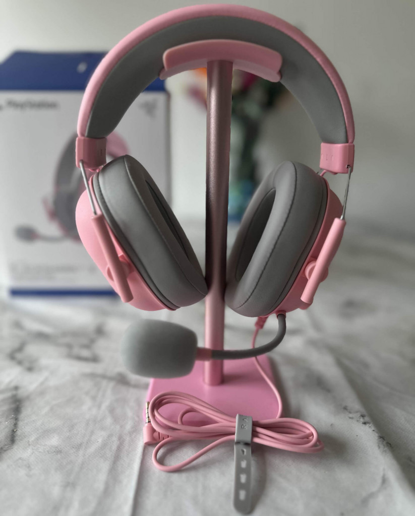 Image of Razer BlackShark V2 X. Pink gaming headset with grey padding on a stand, accompanied by a pink cable, set against a blurred PlayStation box background.