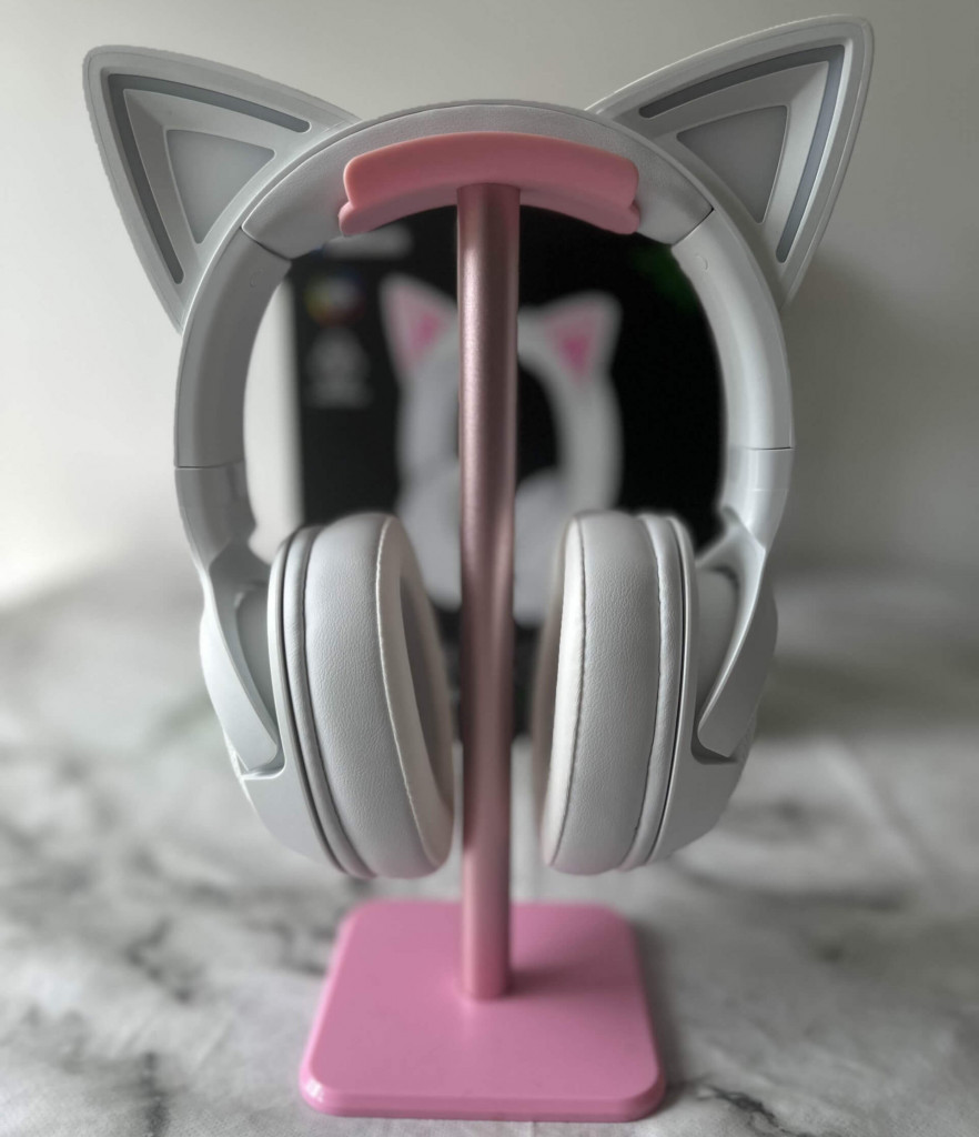 A white Razer Kraken Kitty V2 BT Headset sits on a pink headphone stand on a white marble surface.