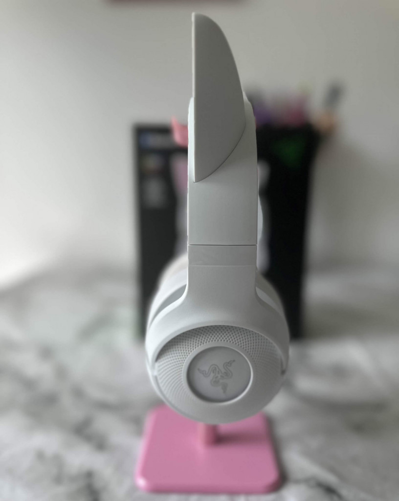 Side on view of a white Razer Kraken Kitty V2 BT Headset sits on a pink headphone stand on a white marble surface.