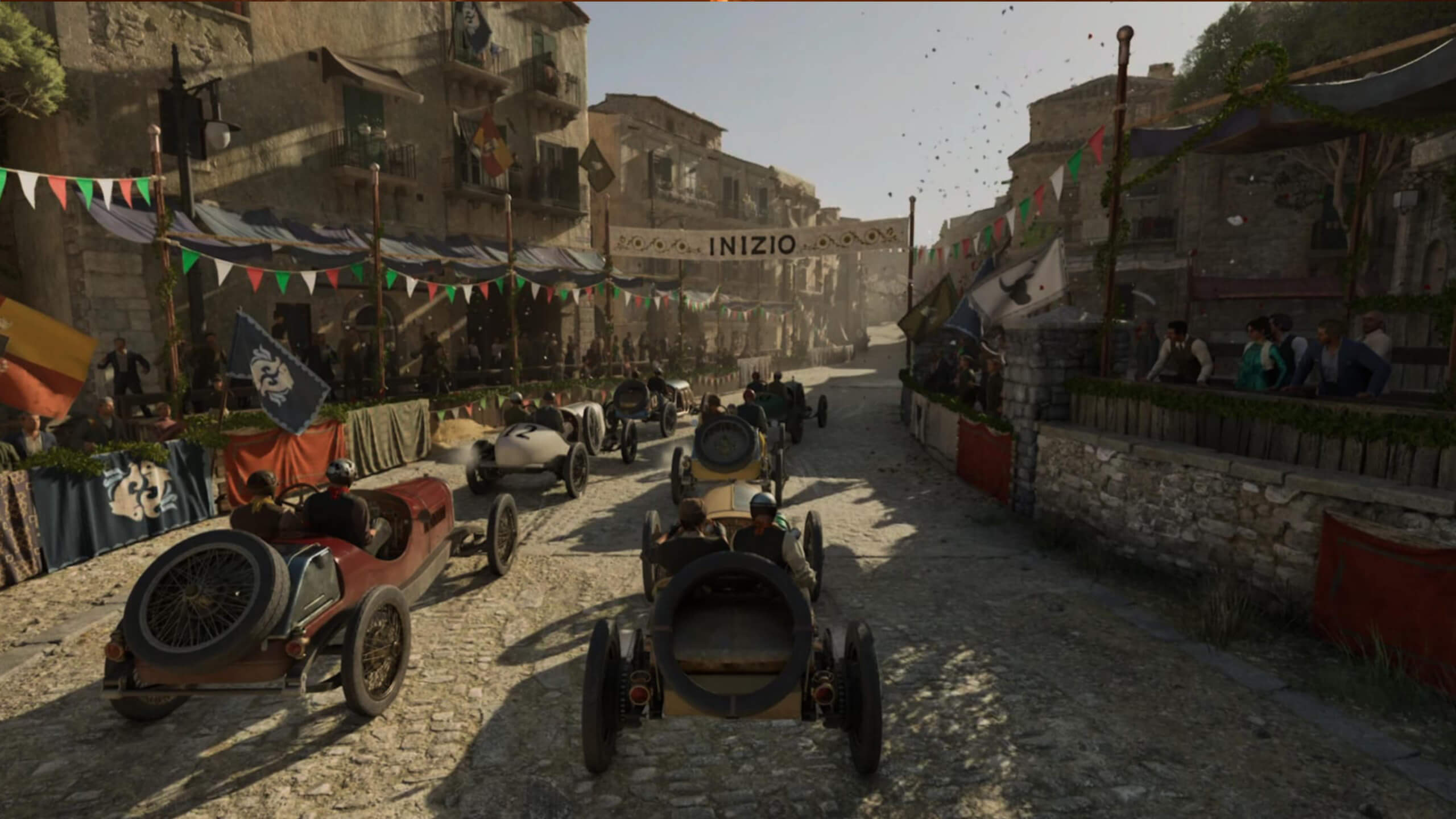 Screenshot from Mafia: The Old Country. Vintage race cars line up on a cobblestone street under a banner reading “Inizio.” Spectators watch from decorated buildings. A lively, historic atmosphere.