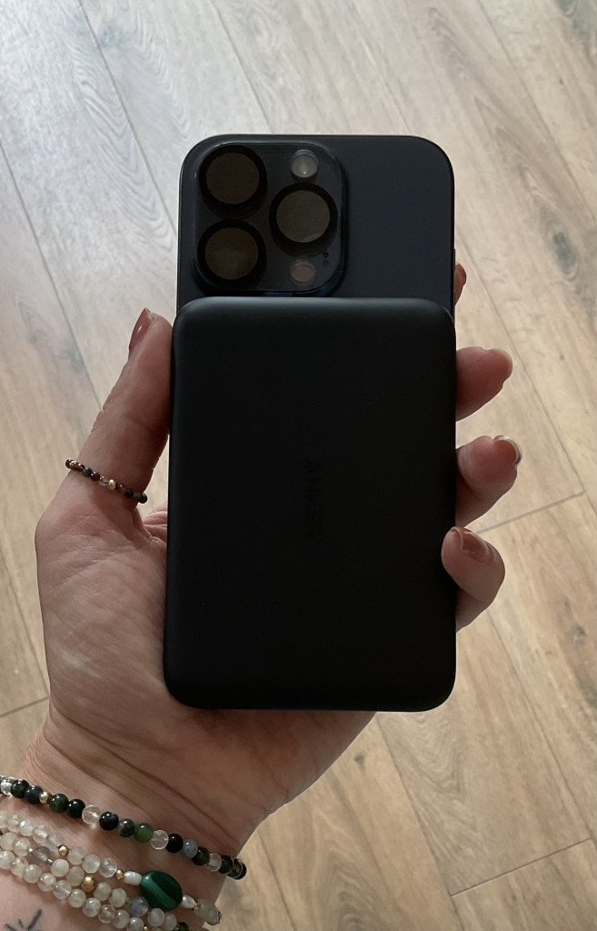 A hand holding a black iPhone with a matching black Anker Nano battery pack attached, surrounded by stone bracelets, on wooden flooring.