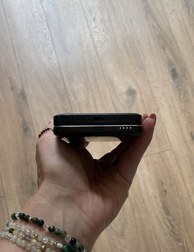 A hand holding a smartphone with an Anker Nano Power Bank stacked, showcasing charging ports and battery indicators on the top device against a wooden floor background.