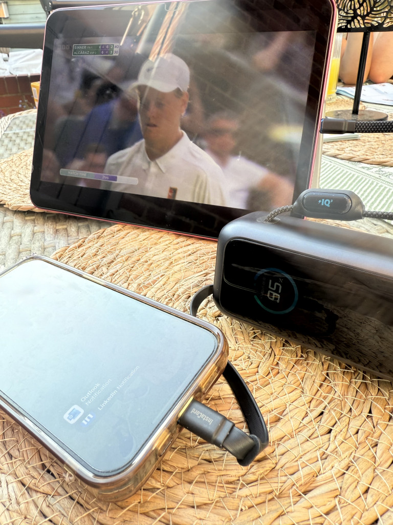 A tablet displaying a sports event sits on a woven placemat, alongside a charging phone and an Anker power bank.
