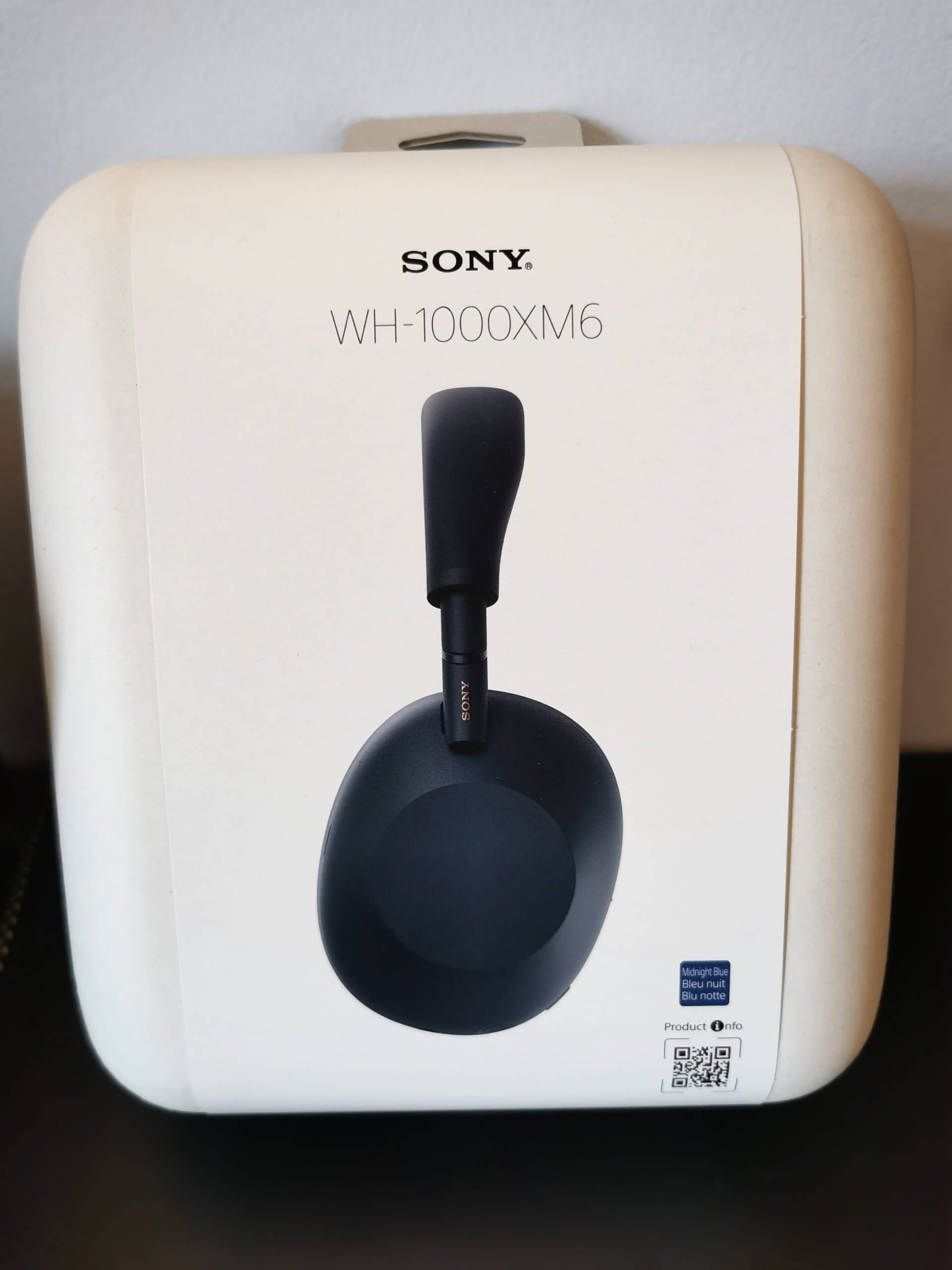 Sony WH-1000XM6 headphones pictured in its white box, lent against a white wall on a black desk.