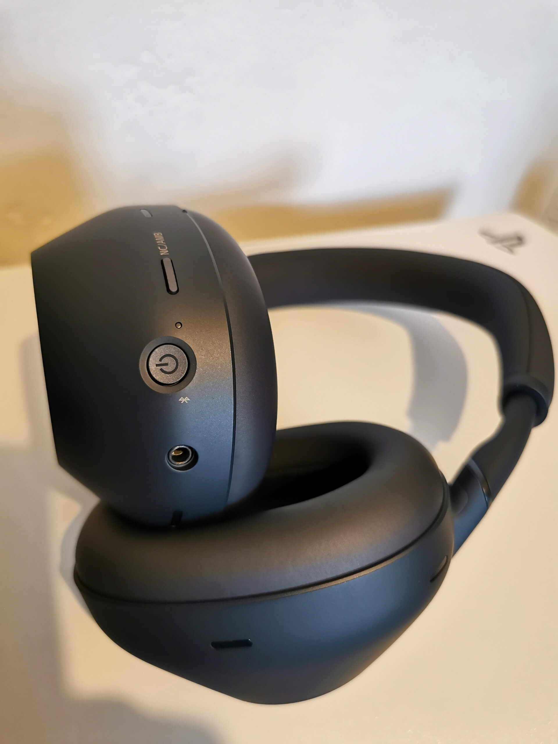 Midnight Blue coloured Sony WH-1000XM6 pictured sat on top of a white PS5. Showing the recessed power button, 3.5mm audio jack, raised NC/AMB button and microphones on the earcups. Also showing the smooth black earpads.