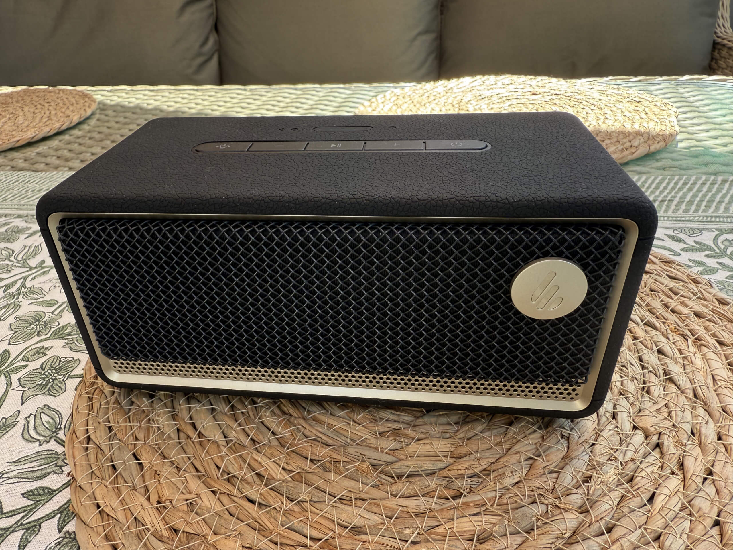 A black portable Bluetooth speaker with a mesh front, metallic accents, and control buttons on top, placed on a woven placemat.