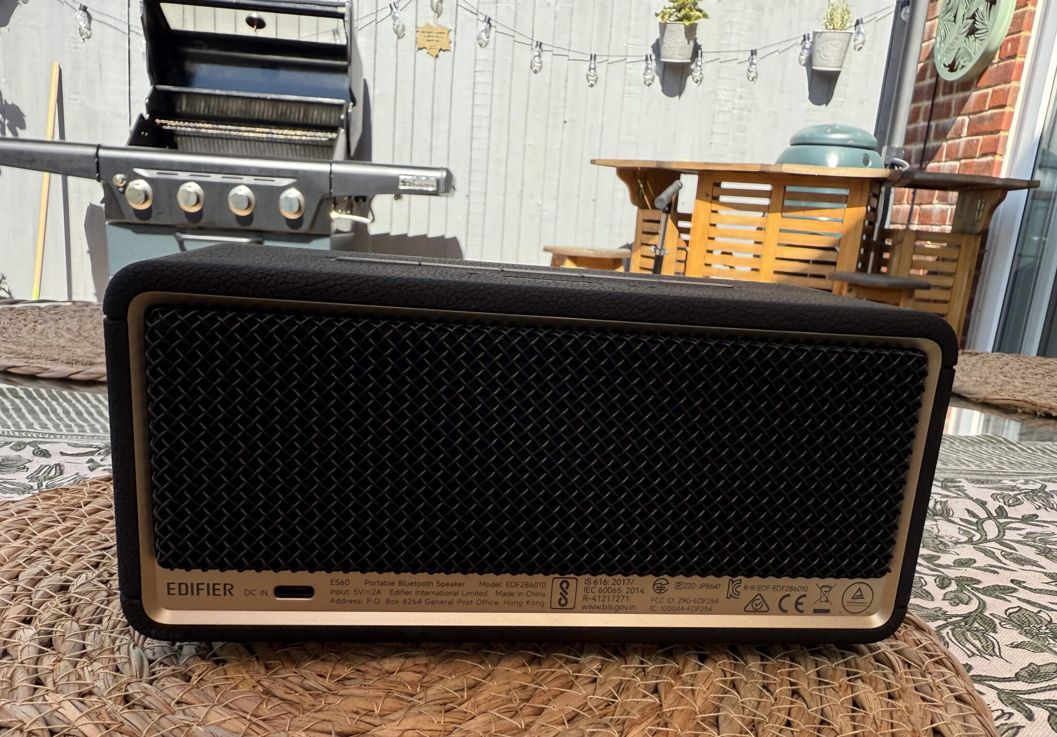 A black portable speaker sits on a woven mat, with a backyard setting featuring a grill and a wooden table in the background. The photo shows the rear of the speaker with the USB C port visible.
