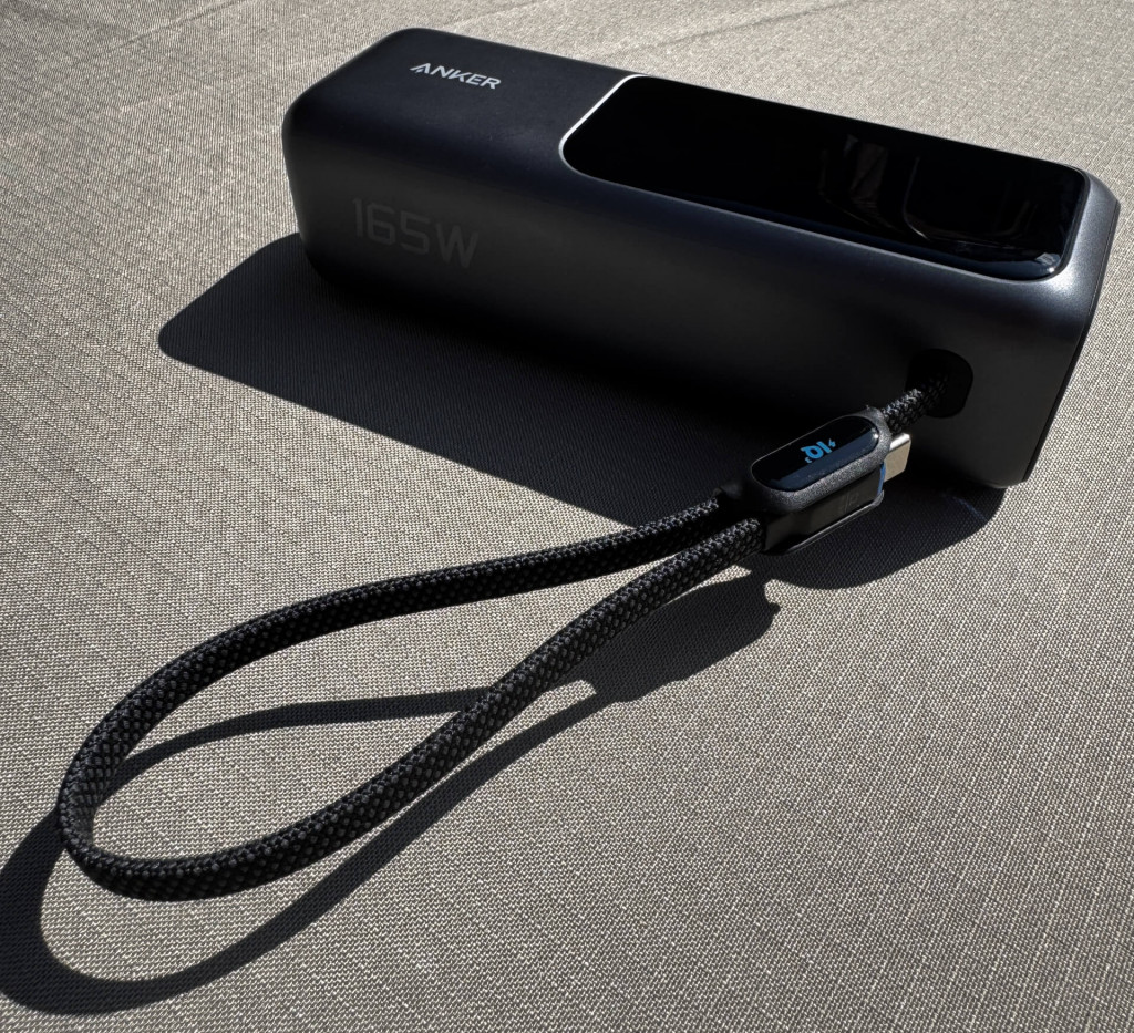 A sleek Anker power bank with 165W capacity, featuring a charging cable loop on a textured surface in sunlight.