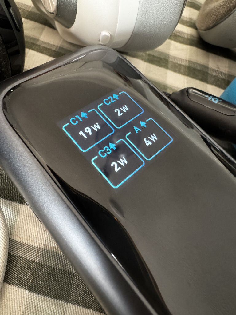 Close-up of an Anker Laptop Power Bank showing power output settings for four ports on a glossy surface, with adjacent headphones visible in the background.