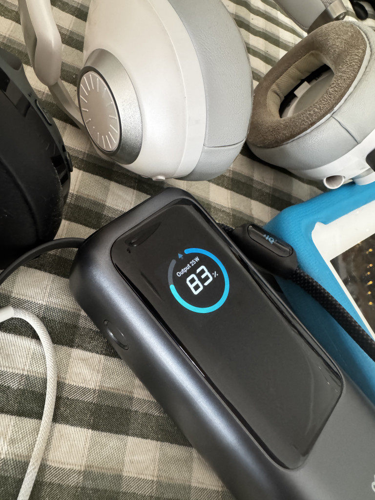 An Anker Laptop power bank displaying an 83% battery charge, surrounded by headphones on a checked fabric surface.
