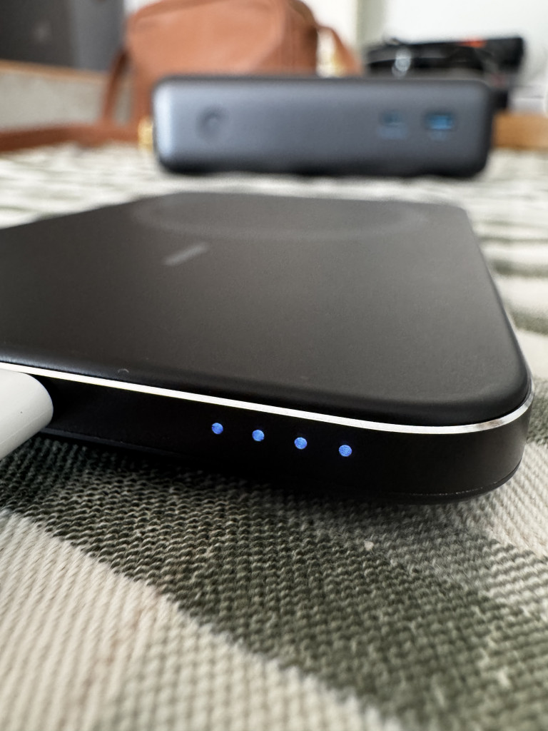 Close-up of a sleek wireless Anker Nano charger with four blue indicator lights, on a checked fabric surface, with a blurred power bank in the background.