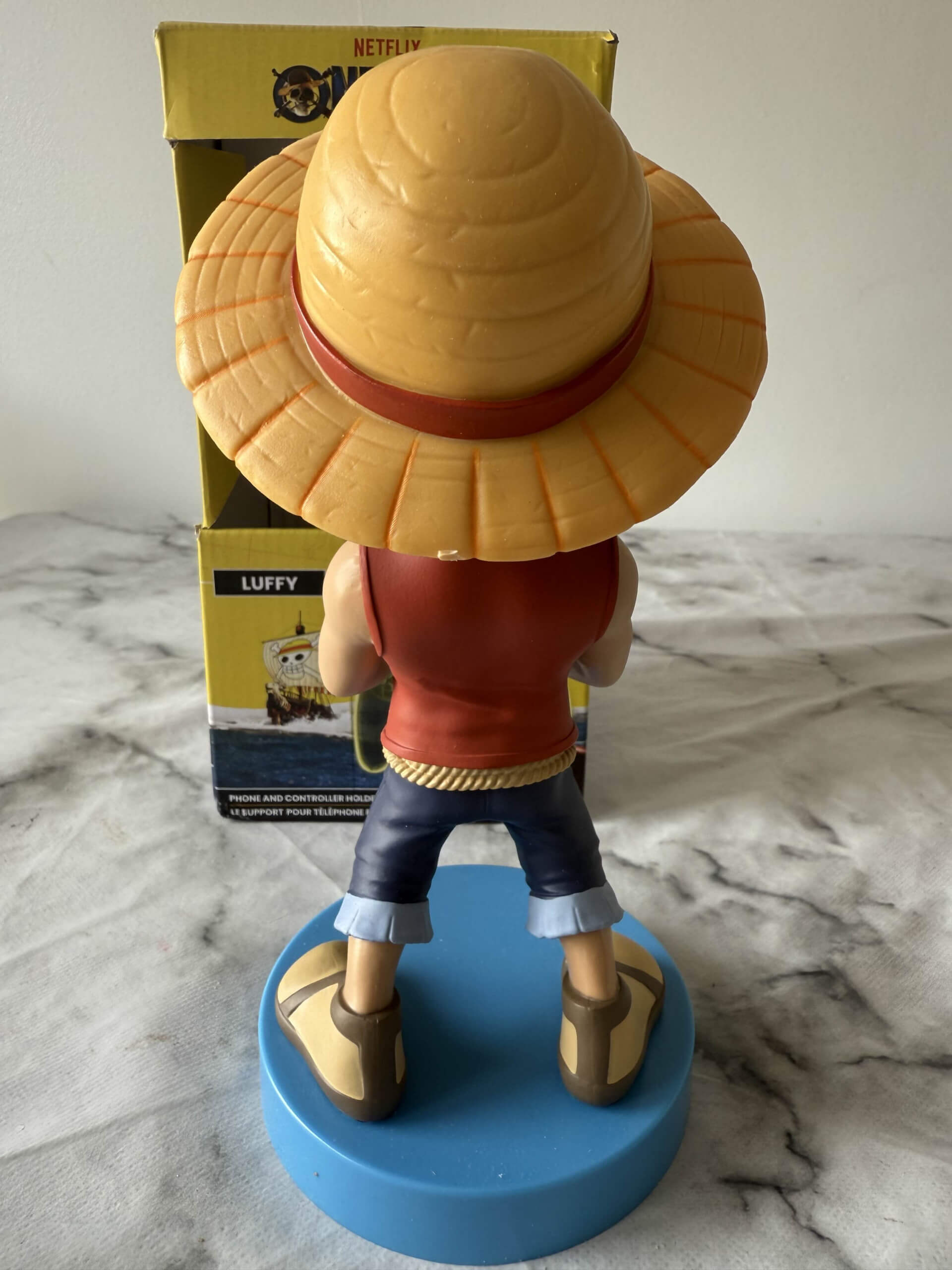 Back of of the Luffy figurine from One Piece, wearing a straw hat and red vest, stands on a blue base.