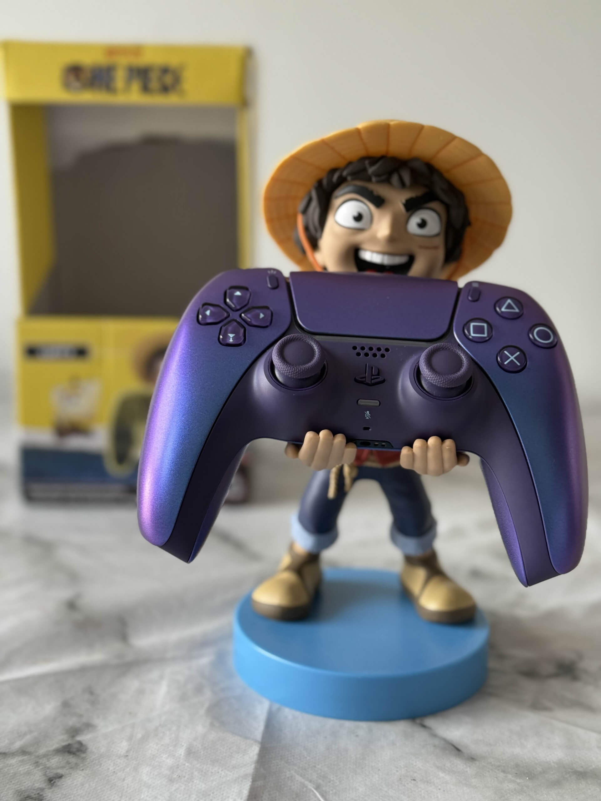 A colourful Luffy Cable Guys figurine holding a purple gaming controller, set against a light background with a yellow box in the background.