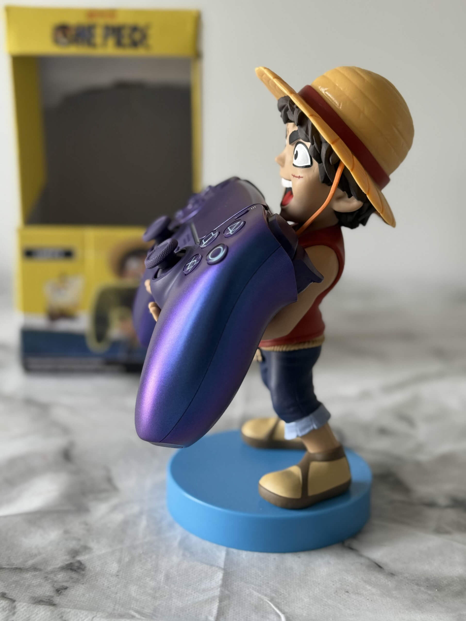 A figurine of a boy in a straw hat joyfully holding a purple game controller, with a yellow box and a blue base in the background.
