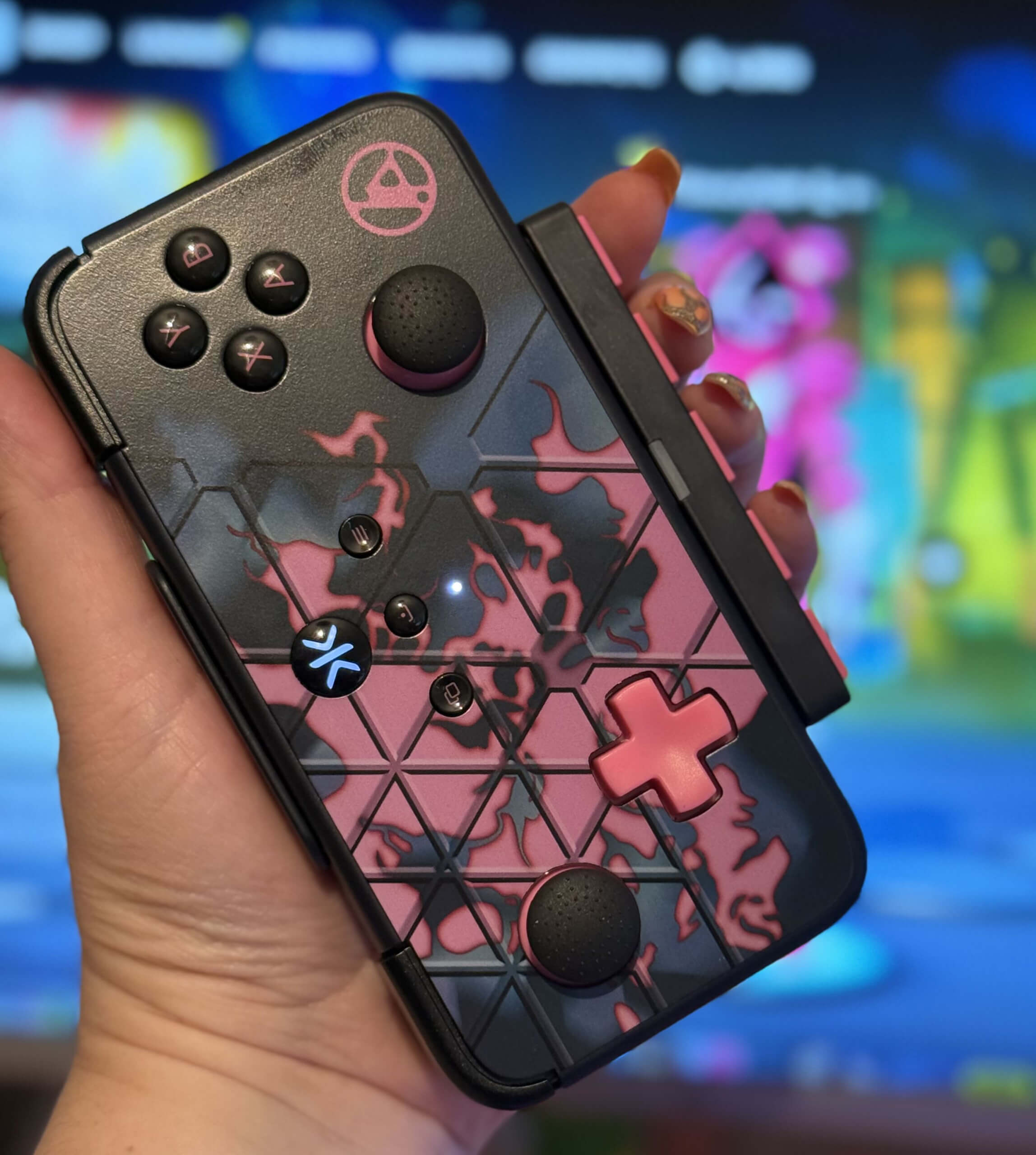 A person holds a neo s linkin park gaming controller with a pink and black flame design against a colourful fortnite festival game background, showing a pink cuddle team leader character blurred.