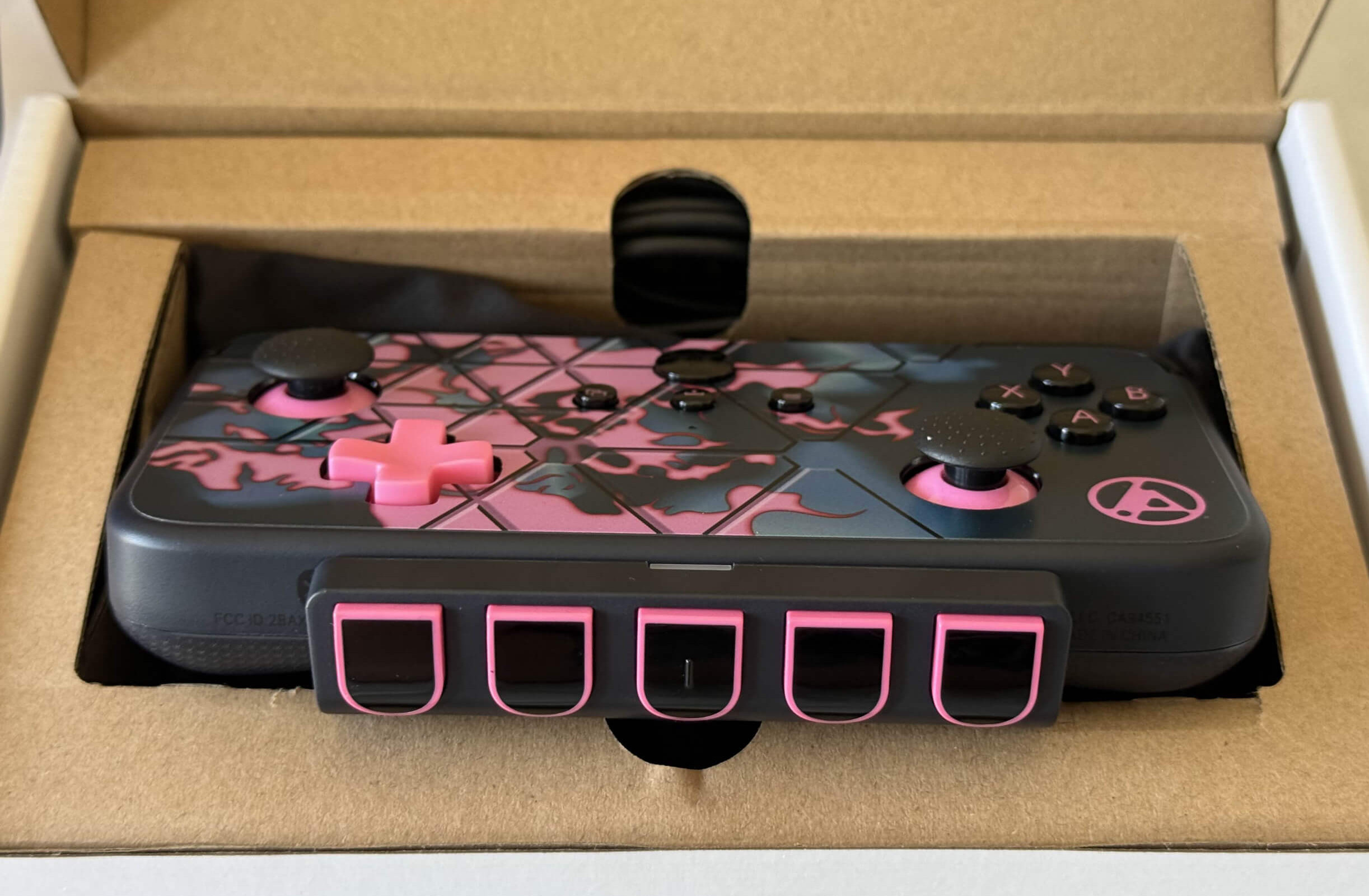 A black and pink gaming controller is displayed in its cardboard packaging, featuring unique design elements and button layout.