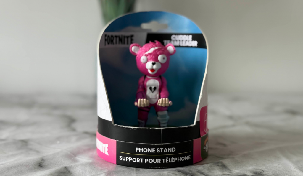 A pink Fortnite Cuddle Team Leader figurine in a packaging designed as a phone stand, set against a blurred background.